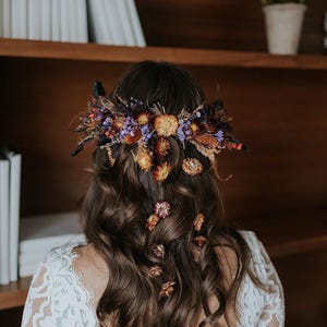 May include: A hair accessory featuring a dense arrangement of dried flowers in shades of orange, purple, and brown, secured at the back of wavy brown hair. Several individual dried flower blossoms are scattered down the length of the hair.