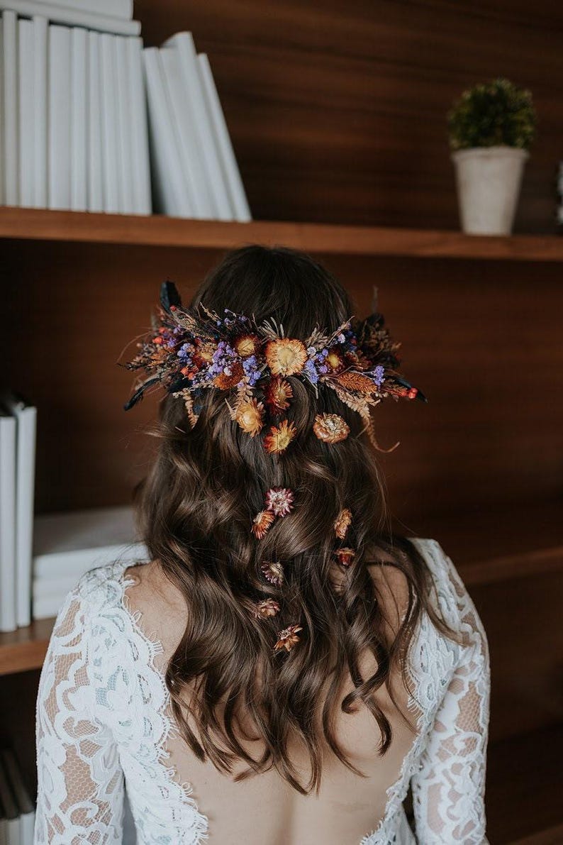 May include: A hair accessory featuring a dense arrangement of dried flowers in shades of orange, purple, and brown, secured at the back of wavy brown hair. Several individual dried flower blossoms are scattered down the length of the hair.