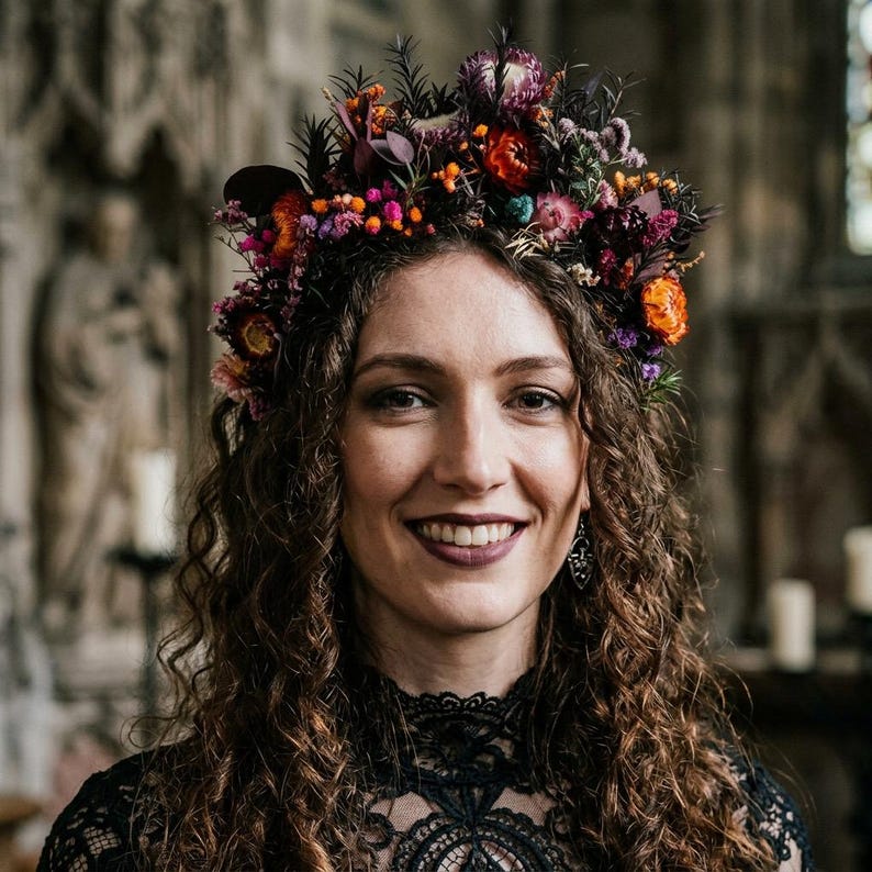 May include: A floral crown featuring a dense arrangement of dried flowers in shades of deep purple, vibrant orange, bright pink, and dark red. The headpiece is composed of various small blossoms, sprigs, and foliage, creating a textured, multi-colored botanical accessory worn atop curly brown hair.
