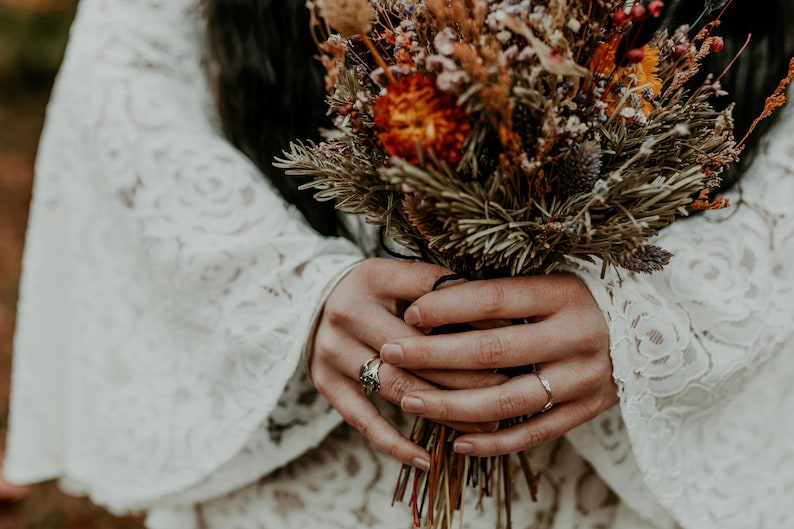 May include: A bouquet of dried flowers in shades of orange, brown, and green, held by hands wearing two silver rings. The bouquet is paired with a white long-sleeved garment featuring an intricate floral lace pattern and wide, flared sleeves.