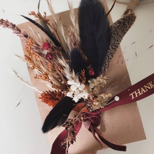 May include: A brown paper gift package adorned with a dried floral arrangement featuring orange and white flowers, tall grasses, and black feathers. A deep red ribbon with the gold text THANK YOU is tied around the package with a small white pearl pin.