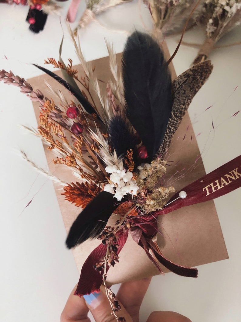May include: A brown paper gift package adorned with a dried floral arrangement featuring orange and white flowers, tall grasses, and black feathers. A deep red ribbon with the gold text THANK YOU is tied around the package with a small white pearl pin.