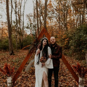 May include: A white lace dress with long bell sleeves and a high leg slit, paired with black boots and a silver crown. The dress features intricate floral patterns and a floor-length hem, worn by a person standing next to a person in a dark blazer.