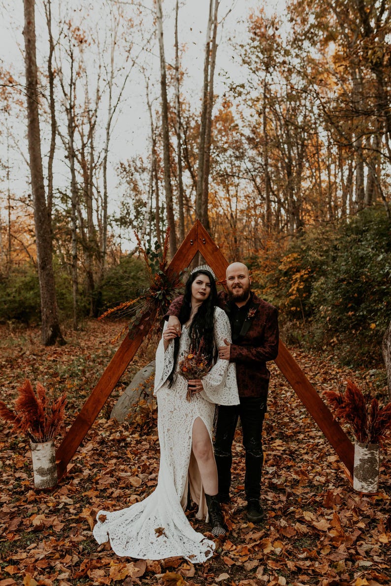 May include: A white lace dress with long bell sleeves and a high leg slit, paired with black boots and a silver crown. The dress features intricate floral patterns and a floor-length hem, worn by a person standing next to a person in a dark blazer.