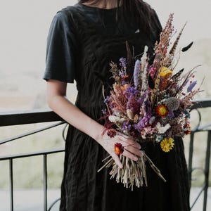 May include: A bouquet of dried flowers featuring a vibrant mix of purple, yellow, orange, and natural brown stems. The arrangement includes various textures, feathery plumes, and small blossoms, held together by a bundle of thin, light brown stalks.