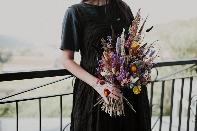May include: A bouquet of dried flowers featuring a vibrant mix of purple, yellow, orange, and natural brown stems. The arrangement includes various textures, feathery plumes, and small blossoms, held together by a bundle of thin, light brown stalks.