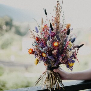 May include: A bouquet of dried flowers featuring a vibrant mix of purple, orange, yellow, and blue stems. The arrangement includes various textures like tall grasses, small round blossoms, and feathery plumes, all tied together at the base with a dark band.