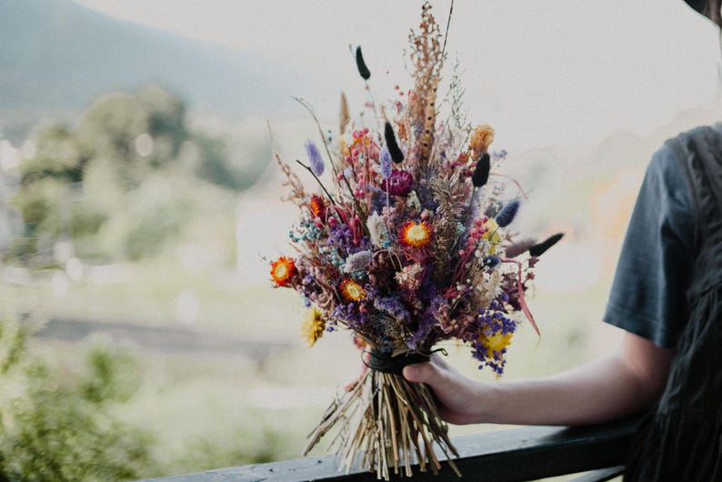 May include: A bouquet of dried flowers featuring a vibrant mix of purple, orange, yellow, and blue stems. The arrangement includes various textures like tall grasses, small round blossoms, and feathery plumes, all tied together at the base with a dark band.