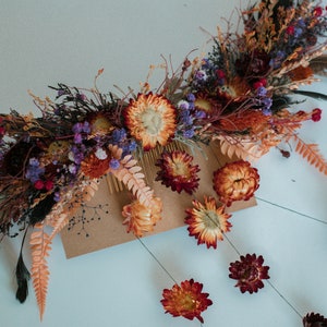 May include: A hair comb adorned with an arrangement of dried flowers and foliage in shades of orange, deep red, and purple. The decorative piece features preserved strawflowers, ferns, and dark feathers, with additional floral stems extending downward.