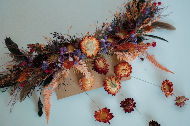 May include: A hair comb adorned with an arrangement of dried flowers and foliage in shades of orange, deep red, and purple. The decorative piece features preserved strawflowers, ferns, and dark feathers, with additional floral stems extending downward.