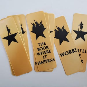 May include: A collection of tan bookmarks featuring black silhouettes of figures and stars. Some bookmarks have text that reads "THE BOOK WHERE IT HAPPENS" and "WORK U'LL E K".