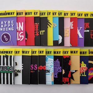 May include: A collection of colorful Broadway-themed magnets. Each magnet features a different show title, with vibrant designs and text. The magnets are rectangular with a yellow top section that reads "BROADWAY".