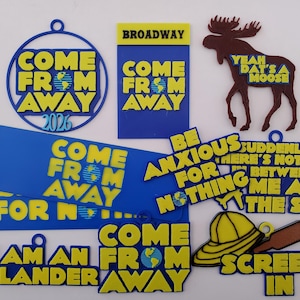 May include: A collection of blue and yellow 3D-printed ornaments and signs featuring text from the musical Come From Away, including phrases like "Be Anxious For Nothing," "I Am An Islander," "Screech In," and a brown moose silhouette.