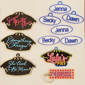 May include: A set of 10 pie-shaped keychains with various sayings, including "Sugar Butter Flour", "Everything Changes", "She Used To Be Mine", and "Lulu's Pies". There is also a keychain with a red and white checkered tag that says "Waitress".