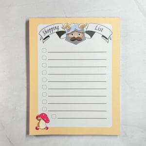May include: A notepad with a light yellow border and a white interior. The top of the notepad has a banner that reads "Shopping List" with a cartoon Viking character. A cartoon mushroom is in the bottom left corner.