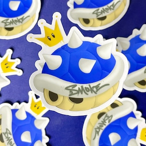 May include: A blue and white spiky shell with a yellow crown on top. The shell has the word "SMOKE" written on it in a graffiti style.