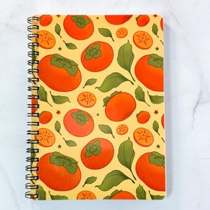 May include: Spiral-bound notebook featuring a repeating pattern of orange persimmons, green leaves, and star shapes on a yellow background. The notebook's binding is black.