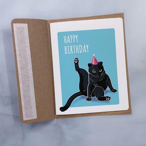 May include: A birthday card featuring a black cat wearing a pink party hat. The cat is sitting on a light blue background with the text "Happy Birthday" in white.