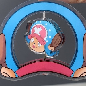 May include: A clear acrylic decoration featuring a cartoon character with a blue hat and a pink cross. The character is inside a blue arch with brown hands on each side and a red base. The character has a surprised expression.