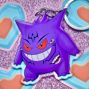 May include: Purple and black Gengar keychain with a menacing grin and red eyes. The keychain is made of clear acrylic and features a silver chain.