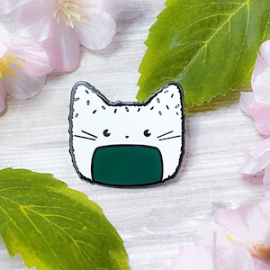 May include: White enamel pin shaped like a cat with a green seaweed filling, resembling a Japanese rice ball, or onigiri. The pin has a white background with black outlines and black dots.