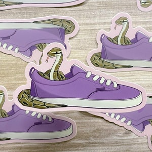 May include: Purple sneakers with a snake wrapped around them. The snake is brown and has a light brown pattern. The sneakers have white laces and a white sole.