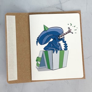 May include: A blue alien character wearing a party hat pops out of a green and white striped gift box. The alien is blowing a red and white striped party favor.