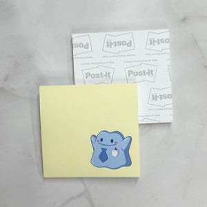 May include: A stack of Post-it notes. The top note is yellow with a blue cartoon character wearing a tie and holding a coffee cup. The Post-it notes are on a white surface.