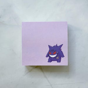 May include: A square, lavender sticky note pad with a purple cartoon character illustration. The character has a wide, toothy grin, red eyes, and spiky hair. The pad is on a white and gray surface.