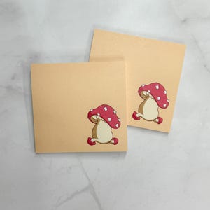 May include: Two square, light tan notecards with a cartoon mushroom illustration. The mushroom has a red cap with white spots and a beige stem. The cards are arranged diagonally on a light-colored surface.