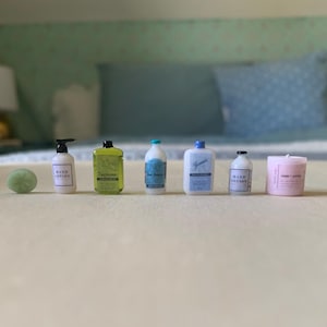 May include: A set of seven miniature bottles and a candle. The bottles are white with black labels and contain hand lotion, hand sanitizer, and body wash. The candle is pink with a white label and says "Rose & Honey".