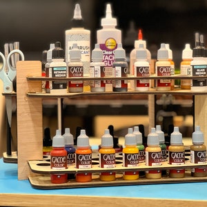 Peut inclure: Un porte-peinture en bois à deux étages contenant divers flacons de peinture, dont des peintures acryliques, un retardateur et un médium de glacis. Les flacons sont disposés en rangées et portent la marque "Game Color".
