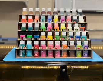 May include: A wooden tiered paint bottle organizer filled with various colors of acrylic paint. The paint bottles are arranged in rows and columns, showcasing a wide spectrum of hues, including pinks, blues, greens, and yellows. Each bottle has a white cap.