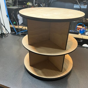 Könnte beinhalten: Ein dreistufiges, drehbares Holzregal mit schwarzem Rand. Jede Stufe hat drei Fächer.