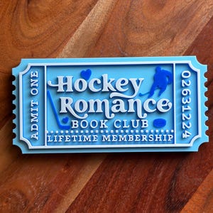 May include: A light blue and white ticket-shaped sign with the words "Hockey Romance Book Club" and "Lifetime Membership". The sign features a hockey player silhouette, a hockey stick, and a puck.