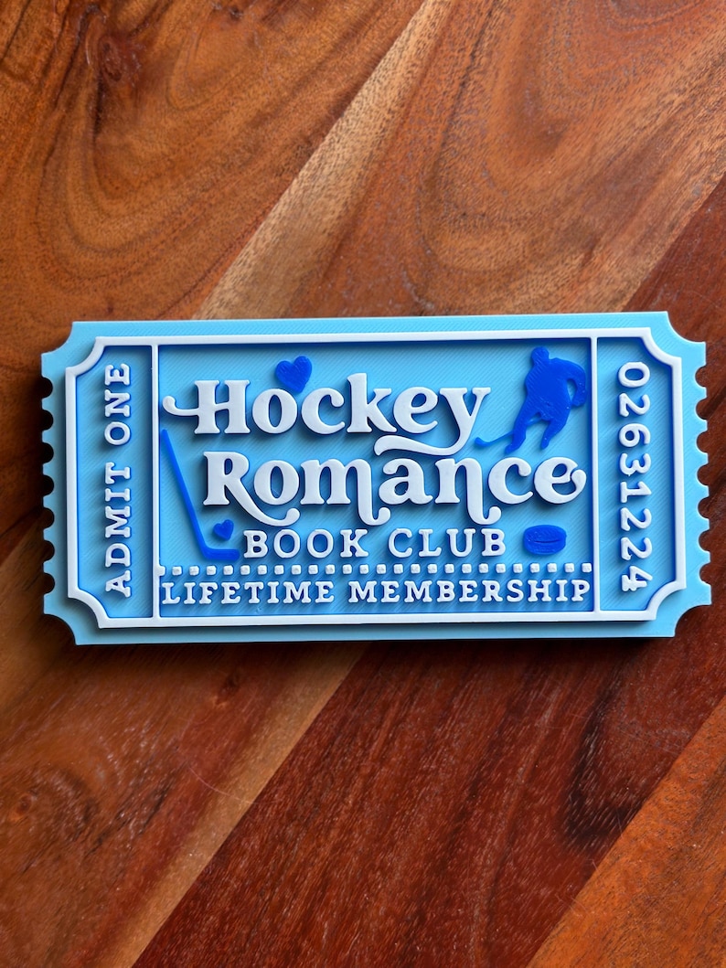 May include: A light blue and white ticket-shaped sign with the words "Hockey Romance Book Club" and "Lifetime Membership". The sign features a hockey player silhouette, a hockey stick, and a puck.