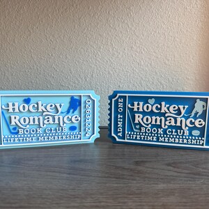 May include: Two decorative tickets, one light blue and one dark blue, with the text "Hockey Romance Book Club Lifetime Membership." Each ticket features a hockey player silhouette and a hockey stick.