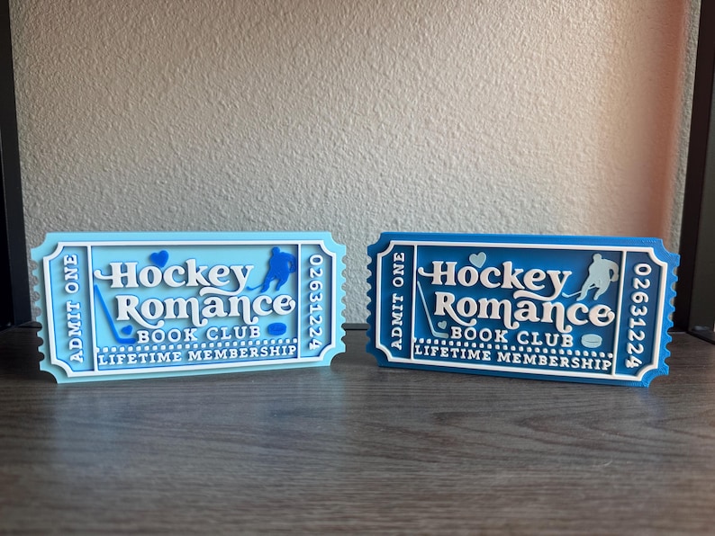 May include: Two decorative tickets, one light blue and one dark blue, with the text "Hockey Romance Book Club Lifetime Membership." Each ticket features a hockey player silhouette and a hockey stick.