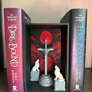 May include: A bookshelf display featuring two books titled "The Wolves of Ruin" with "Dire Bound" and "Fury Bound" on the spines. A decorative scene between the books includes a sword, a red moon, and two white wolf figurines.