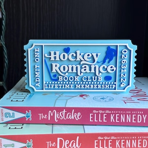 May include: A light blue ticket-shaped sign that reads "Hockey Romance Book Club Lifetime Membership" with a hockey player graphic. The sign is placed on top of two books titled "The Mistake" and "The Deal" by Elle Kennedy.