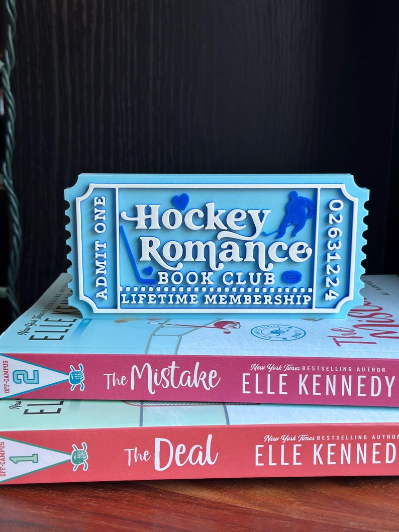 May include: A light blue ticket-shaped sign that reads "Hockey Romance Book Club Lifetime Membership" with a hockey player graphic. The sign is placed on top of two books titled "The Mistake" and "The Deal" by Elle Kennedy.