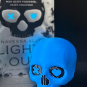 May include: A blue skull-shaped decorative object with black eye sockets, one with a cross and the other with a heart. A book with a skull design and the title "LIGHT OUT" is in the background.