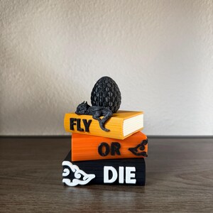 May include: A stack of three rectangular blocks resembling books with the text FLY OR DIE printed on the spines. A black textured dragon egg and a small black dragon figure rest on top of the yellow book. The middle book is orange and the bottom book is black with white cloud-like symbols.