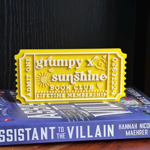 Puede incluir: Un letrero amarillo y blanco con forma de boleto que dice "grumpy x sunshine BOOK CLUB LIFETIME MEMBERSHIP". El letrero tiene un dise&ntilde;o de sol y coraz&oacute;n. El boleto est&aacute; colocado encima de un libro con el t&iacute;tulo "Assistant to the Villain".