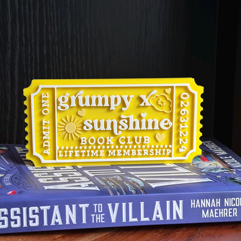 Puede incluir: Un letrero amarillo y blanco con forma de boleto que dice "grumpy x sunshine BOOK CLUB LIFETIME MEMBERSHIP". El letrero tiene un dise&ntilde;o de sol y coraz&oacute;n. El boleto est&aacute; colocado encima de un libro con el t&iacute;tulo "Assistant to the Villain".