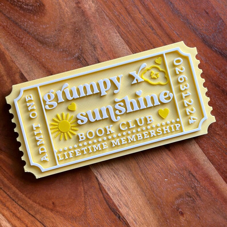 Puede incluir: Un objeto amarillo y blanco con forma de boleto con las palabras "Grumpy x Sunshine Book Club Lifetime Membership". El dise&ntilde;o incluye un sol, corazones y una nube. El boleto dice "Admit One" y tiene un n&uacute;mero de serie.