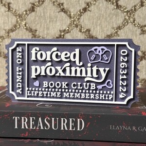 May include: A layered, ticket-shaped sign with the words "forced proximity BOOK CLUB LIFETIME MEMBERSHIP". The sign is dark gray with white and light purple accents. The sign is placed on top of two books with the titles "TREASURED" and "TORMENTED".
