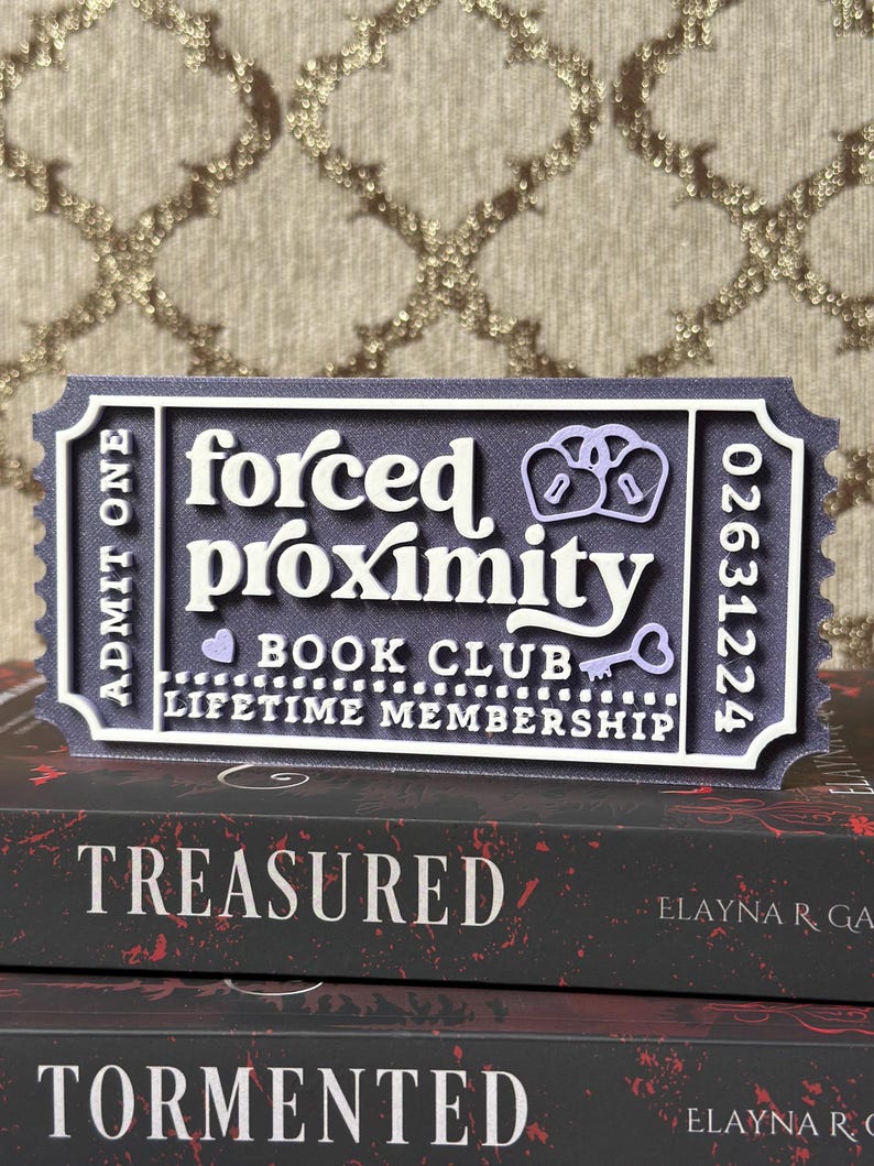 May include: A layered, ticket-shaped sign with the words "forced proximity BOOK CLUB LIFETIME MEMBERSHIP". The sign is dark gray with white and light purple accents. The sign is placed on top of two books with the titles "TREASURED" and "TORMENTED".