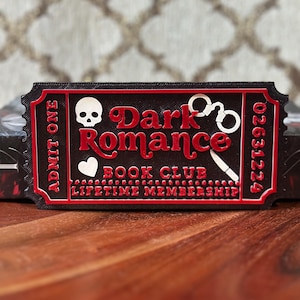 Puede incluir: Un objeto en forma de boleto negro y rojo con las palabras "Dark Romance Book Club Lifetime Membership". El boleto presenta una calavera, un coraz&oacute;n, esposas y un cuchillo. Tambi&eacute;n es visible el texto "Admit One" y un n&uacute;mero de serie.