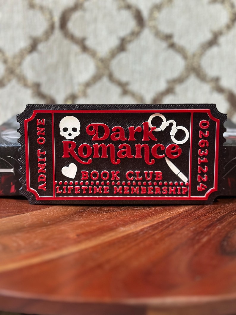 Puede incluir: Un objeto en forma de boleto negro y rojo con las palabras "Dark Romance Book Club Lifetime Membership". El boleto presenta una calavera, un coraz&oacute;n, esposas y un cuchillo. Tambi&eacute;n es visible el texto "Admit One" y un n&uacute;mero de serie.